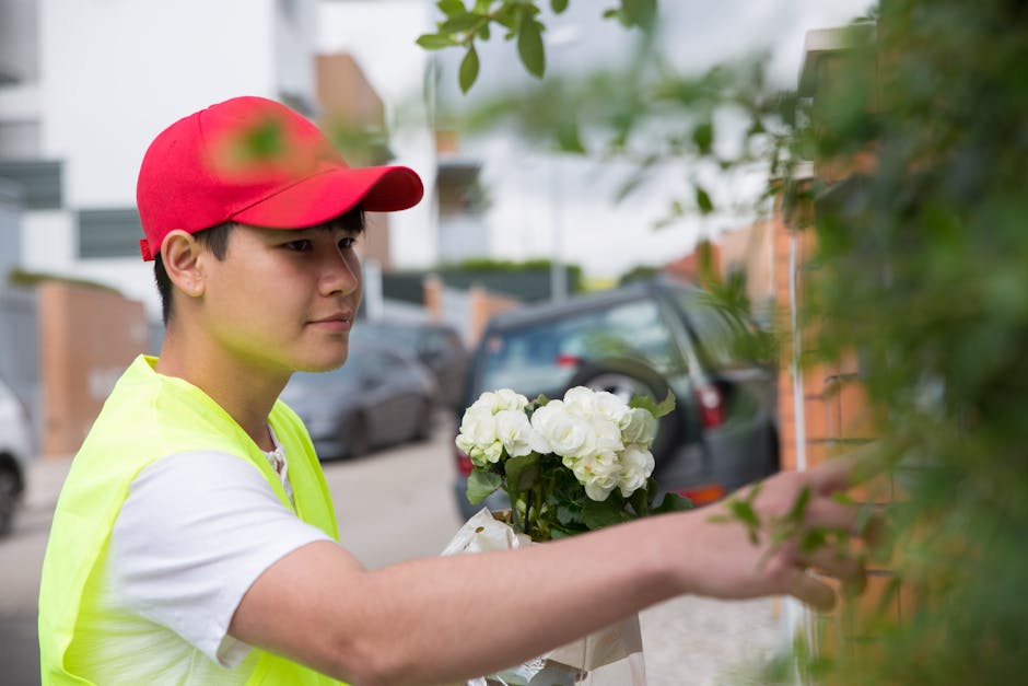 A young male florist wearing a red cap and a neon yellow vest arranging a bouquet of white hydrangeas outdoors during daylight. The bouquet is wrapped in clear plastic, and the florist is placing the 