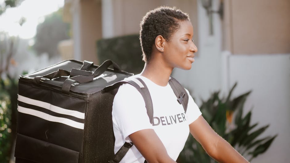 A young woman with short, textured hair, wearing a white T-shirt with the word 'DELIVERY' printed on it, is standing outdoors. She is holding a large, black insulated delivery bag with white stripes o