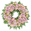 In this softly lit photograph, a round sympathy wreath is laid flat on a spotless white background, its circular shape symbolising continuity and gentle remembrance. The wreath is richly filled with roses in gracefully subdued hues of dusty lavender, antique mauve, and greyish purple, each bloom fully open to reveal layers of velvety petals curling into a soft centre. Nestled among these larger roses are clusters of blush pink spray roses and small rosebuds at different stages of opening, from tight buds to partially unfurled blossoms. Their fresh pastel pink introduces a delicate brightness that softens the deeper tones, adding tenderness and textural interest. The blooms are supported by a dense ring of dark green fern fronds, likely leatherleaf or sword fern, whose finely cut, serrated leaves spread outwards beneath the flowers. This luxuriant foliage frames the wreath like a living halo, its deep green echoing the urban greenery and riverside planting to be found near Deptford's streets and parks. Small hints of darker filler greenery and tiny buds tucked between the roses contribute depth and dimension, so the wreath appears full and layered, not flat. The overall composition is carefully balanced and symmetrical, with each flower thoughtfully placed to maintain harmony around the circle. The atmosphere is one of quiet respect and consolation, making this wreath an appropriate choice for a funeral service in Deptford, a memorial at a local church, or a graveside tribute. It expresses sympathy and enduring love through its soft colour palette, lush texture, and evident craftsmanship.